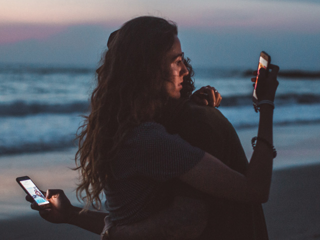 Pareja revisando cada una por su cuenta el móvil, mientras se abrazan en la playa